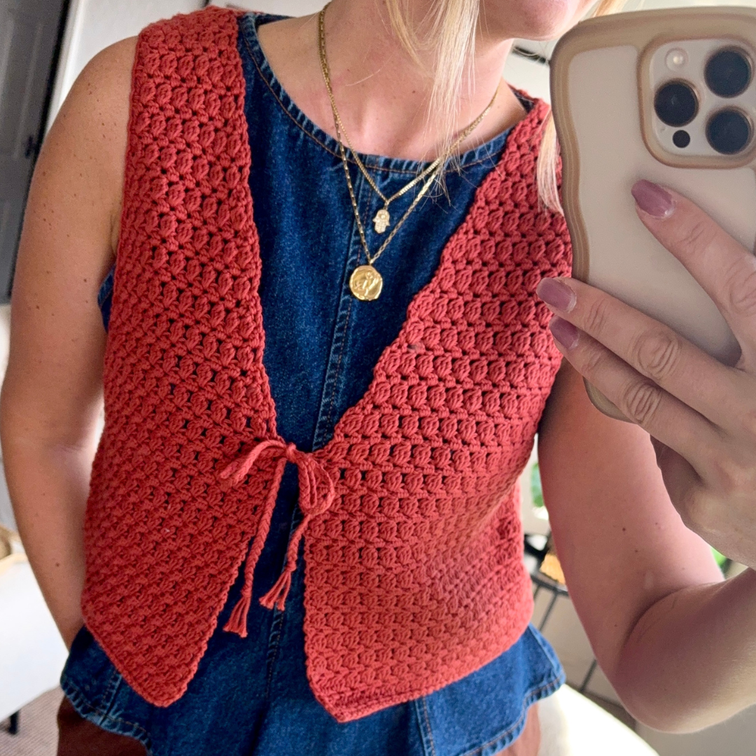Person wearing a red crochet vest over a blue dress, holding a phone.